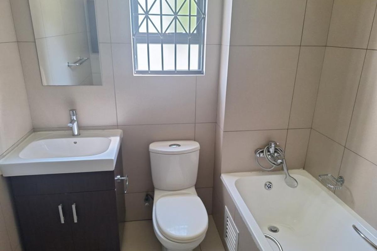 A clean, modern bathroom featuring a vanity cabinet, toilet, and bathtub with tiled walls and a window with security bars.