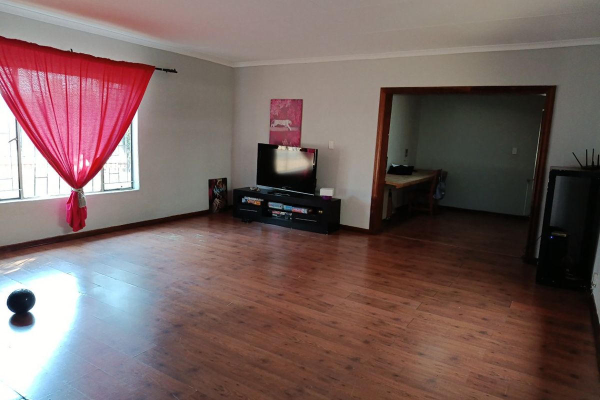 Spacious living room featuring warm-toned laminate flooring and a large window with red curtains, leading into an adjacent room through a timber-framed opening.