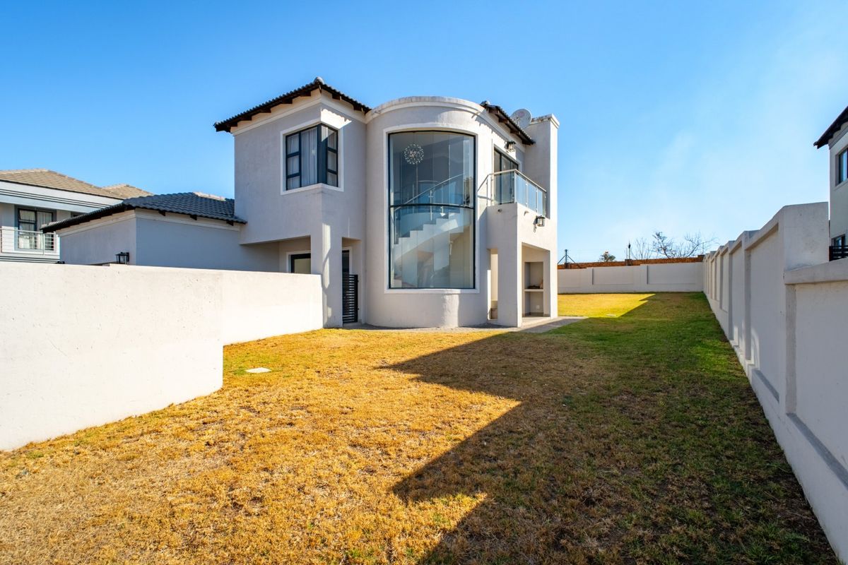 Rear view of a modern double-story home featuring a distinctive curved glass facade, a balcony, and a built-in braai area overlooking the garden.