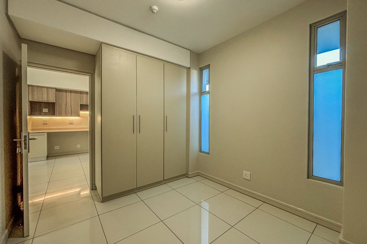 A clean, empty bedroom featuring modern tiled flooring, built-in cupboards, and vertical windows, with a view into an adjacent kitchen area.