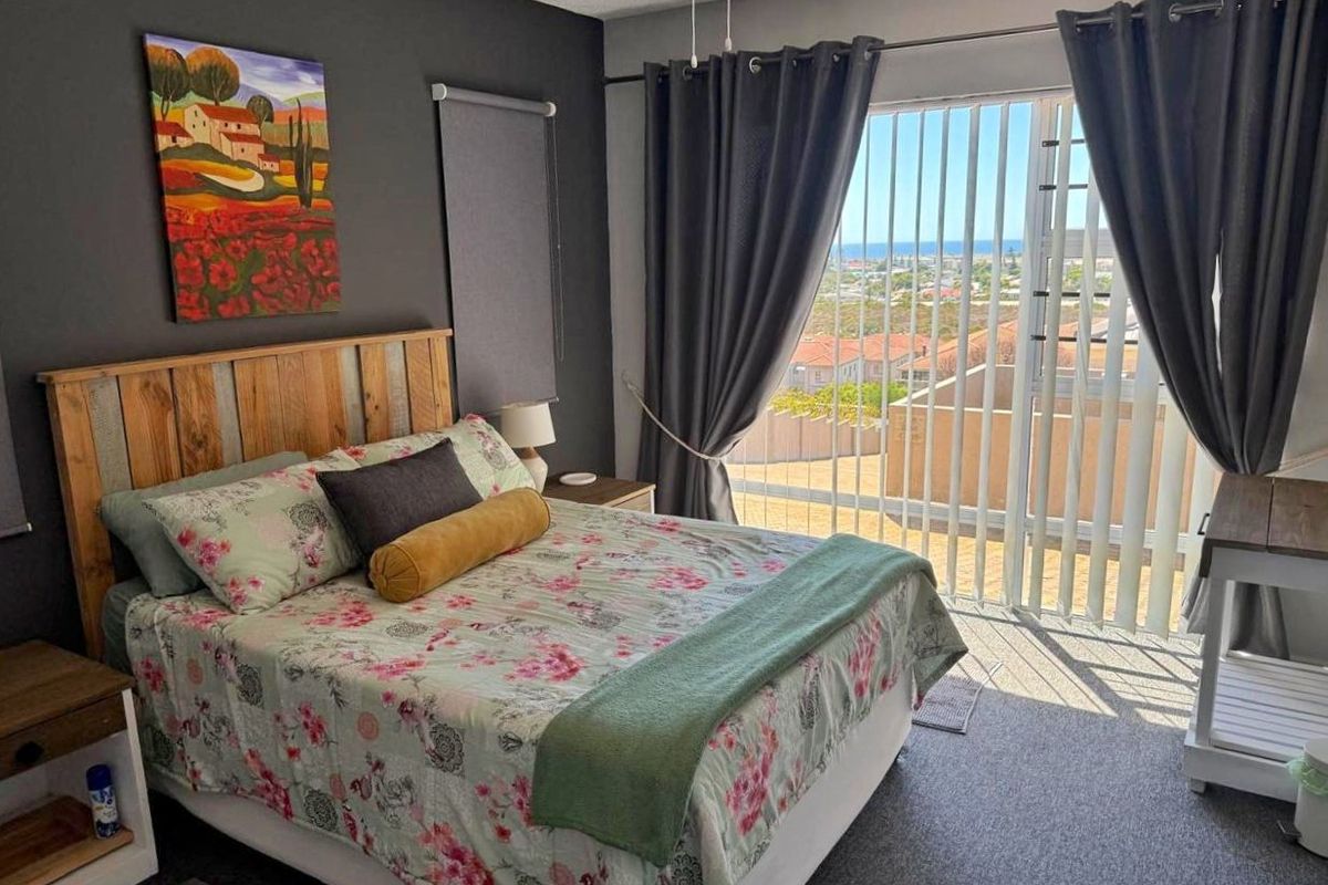 A well-lit bedroom featuring carpet flooring, a ceiling fan, and a sliding glass door providing access to a balcony with coastal views.