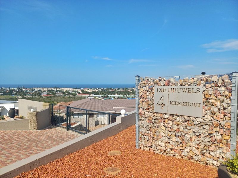 Front entrance area featuring a modern gabion wall, paved driveway, and elevated views of the surrounding neighborhood and coastline.
