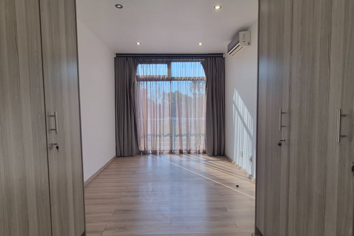 A bright, empty bedroom featuring modern laminate flooring, extensive built-in cupboards, recessed lighting, and an air conditioning unit with access to a balcony.