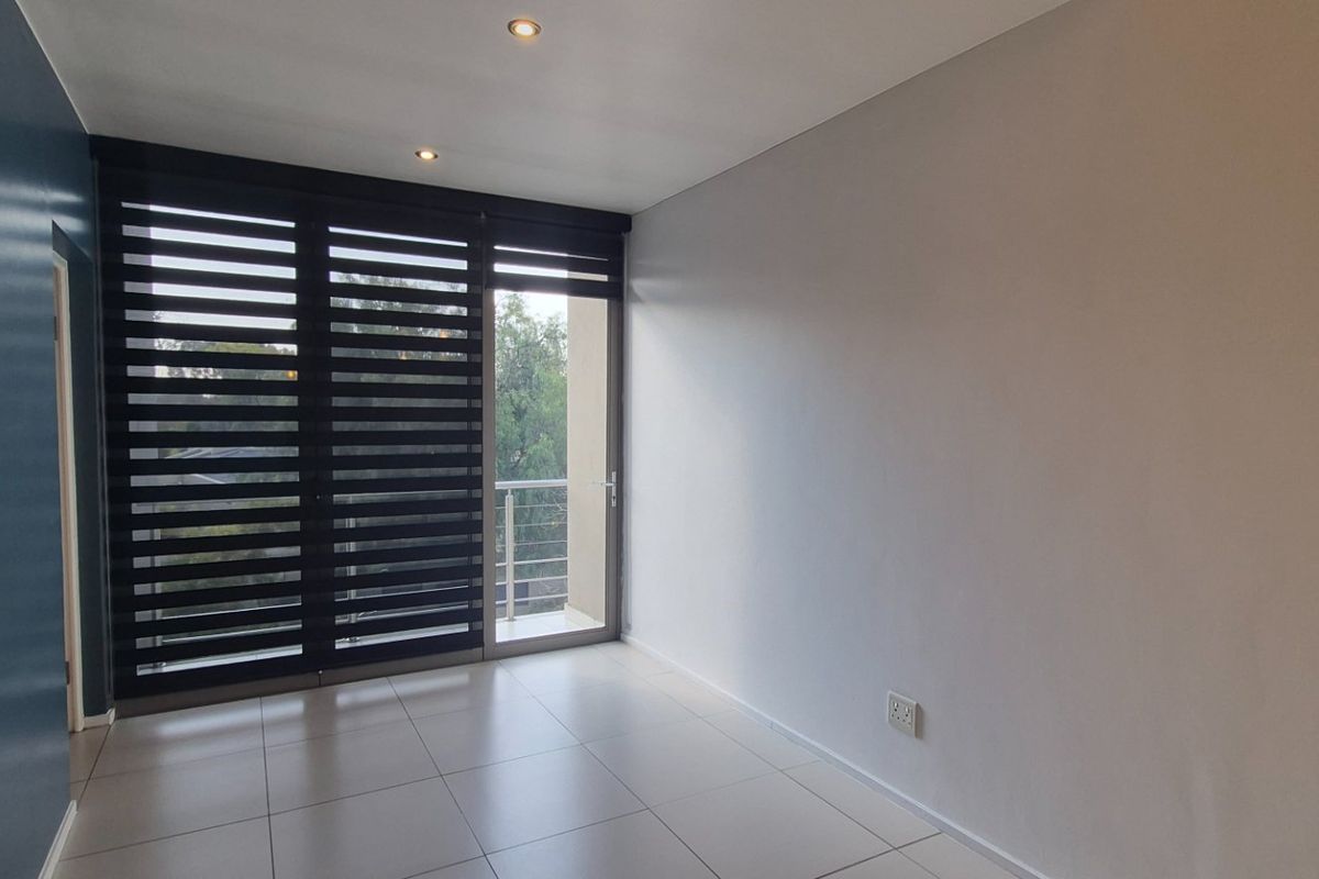 A bright, empty living area featuring neutral tiled flooring, recessed lighting, and a large sliding glass door with modern roller blinds leading to a balcony.