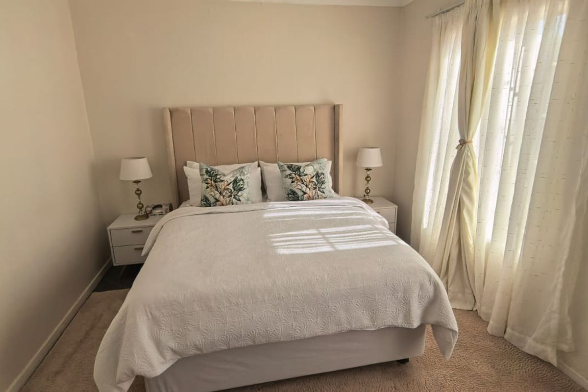 A well-maintained bedroom featuring neutral wall tones, carpeted flooring, and large windows dressed with light curtains.