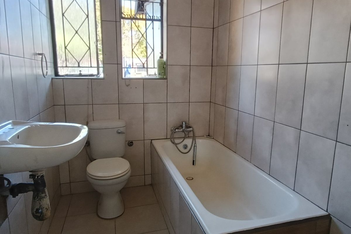 A functional bathroom featuring a built-in bathtub, pedestal washbasin, and toilet, finished with full-height ceramic wall tiling and security bars on the window.