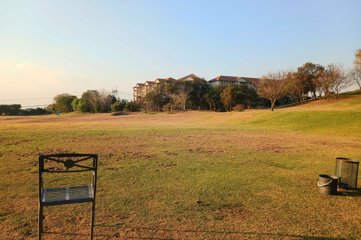 A wide, open grassy area featuring established trees and a view of neighboring residential buildings.