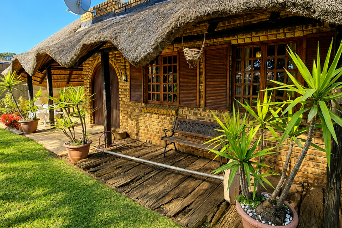 Rear exterior view of a rustic face brick house featuring a traditional thatch roof, timber window shutters, and a wooden deck area.