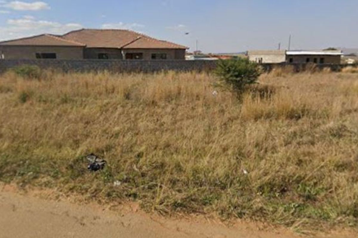 A view of a vacant residential plot featuring overgrown grass and a perimeter wall, with neighboring structures visible in the background.