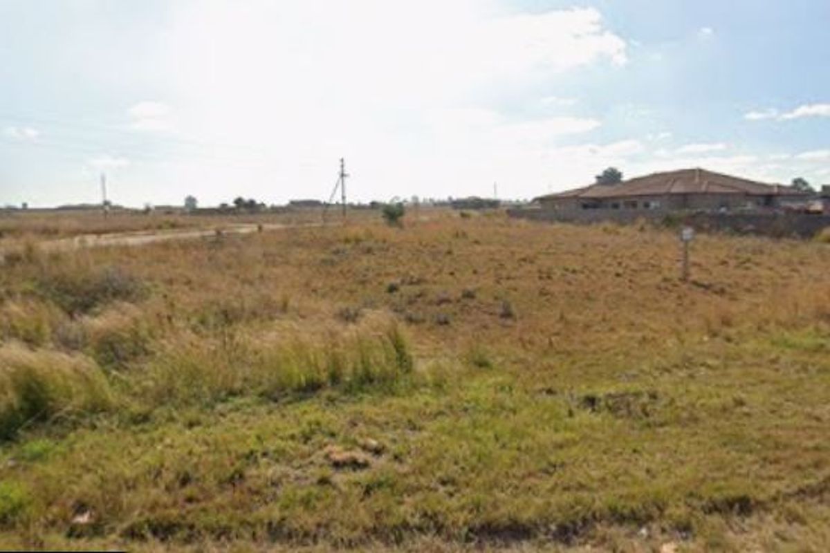A vacant residential plot of land featuring natural grass cover, adjacent to an unpaved road with nearby residential development.