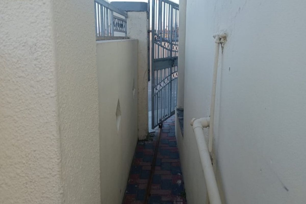 A narrow side passage featuring a paved walkway, boundary wall, and metal security gate, with visible external plumbing infrastructure.
