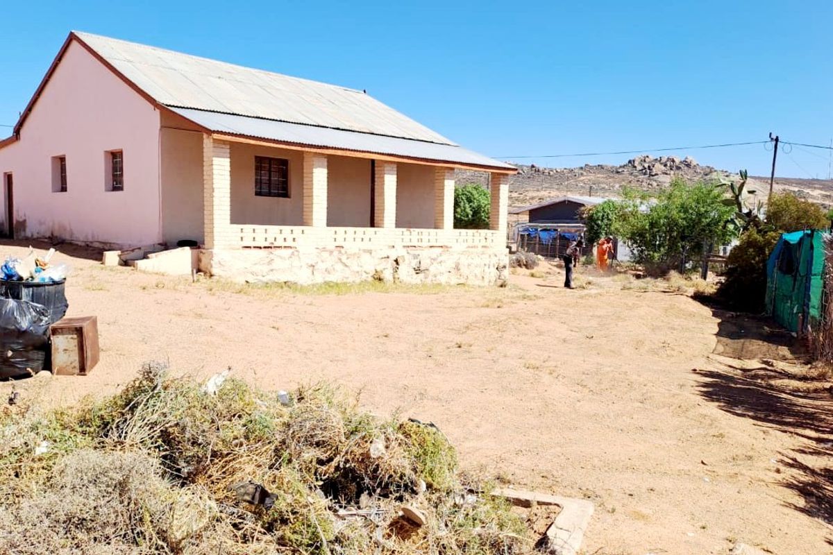 Rear view of a single-story house featuring a covered patio with brick pillars, a corrugated metal roof, and a stone foundation on a large plot.