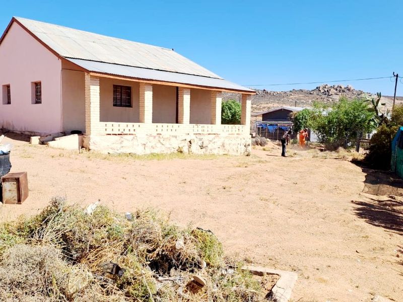 Rear view of a single-story house featuring a covered patio with brick pillars, a corrugated metal roof, and a stone foundation on a large plot.