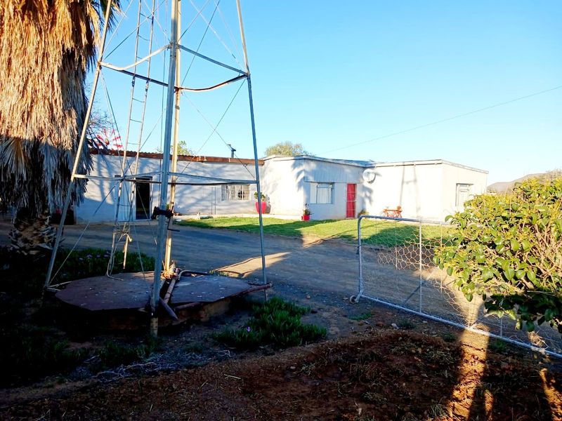 Front view of a farm property featuring a traditional windmill, gravel driveway, and single-story dwelling with a metal security gate.