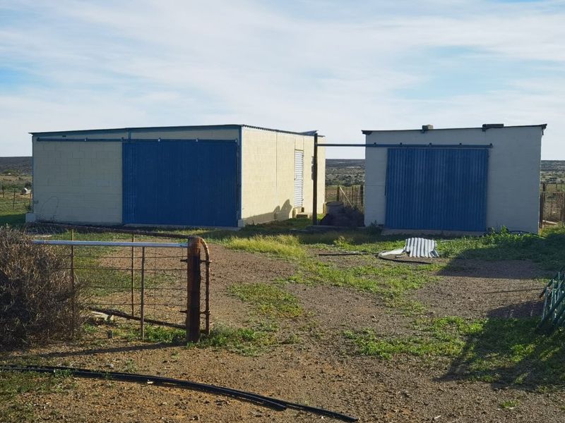 Two detached utility or storage structures with sliding metal doors, situated on a rural farm property with gravel and grass surroundings.