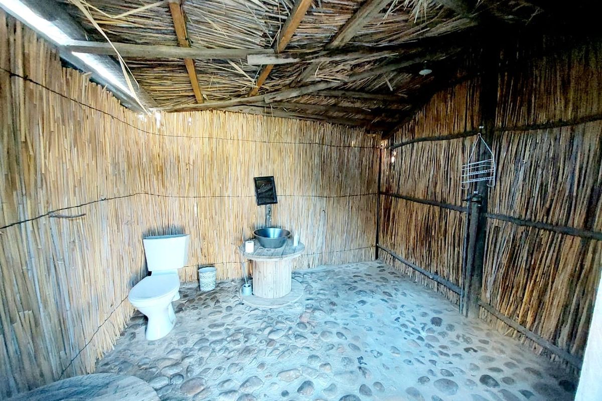 Rustic outdoor-style bathroom featuring a stone floor, reed wall cladding, thatched roof, toilet, and a vessel sink on a wooden vanity.