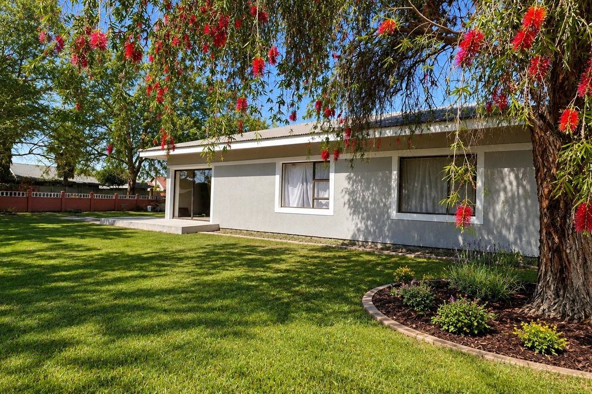 A well-maintained rear exterior view of the house featuring a concrete patio, expansive lawn, and established garden beds.