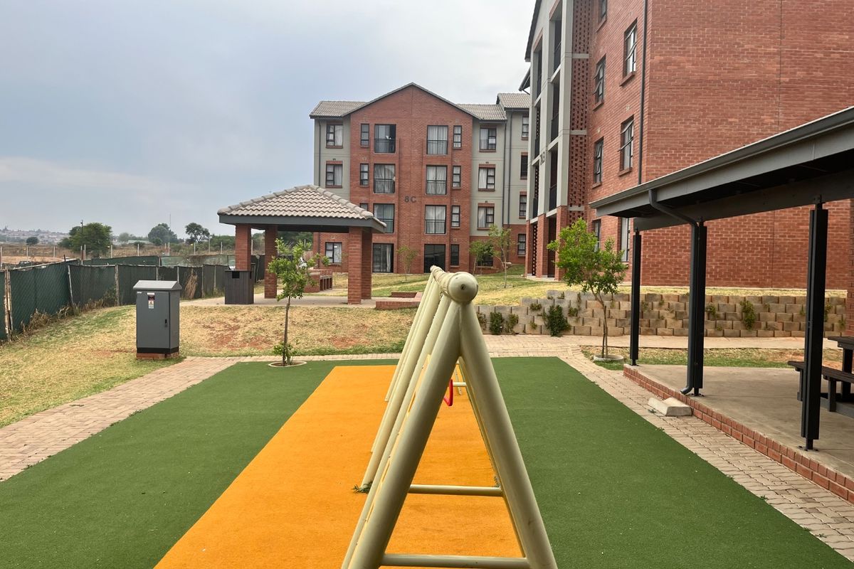 Communal outdoor area featuring a brick apartment facade, covered patio, gazebo, and a playground with synthetic turf surfacing.