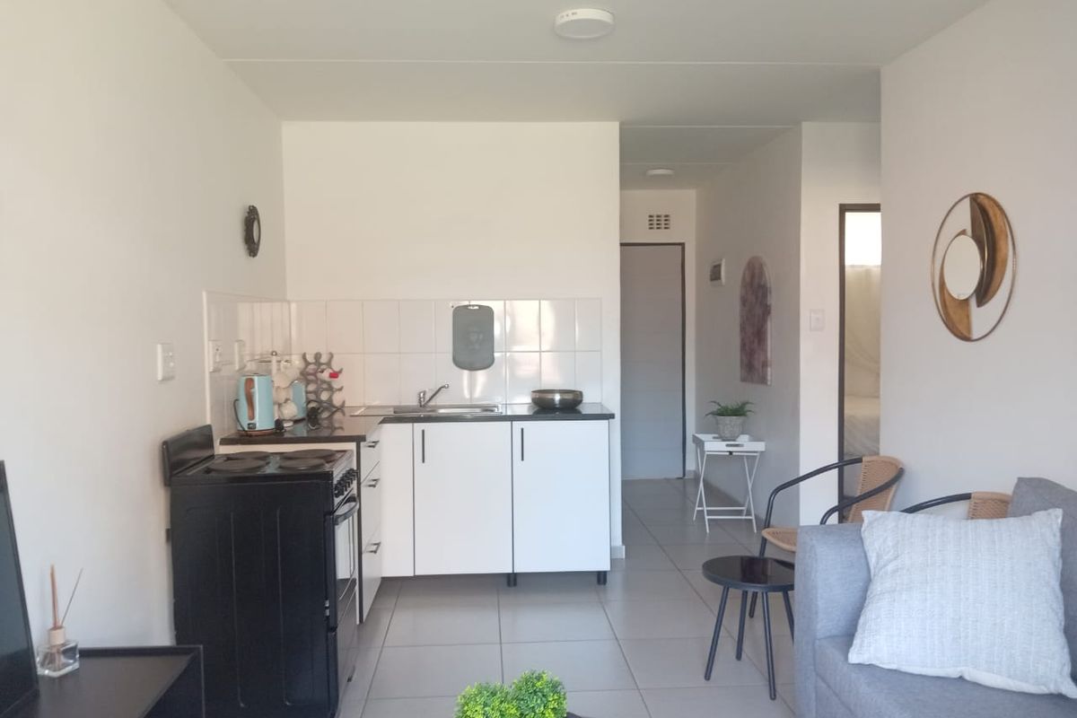 Open-plan living area featuring a compact kitchen with white cabinetry, tiled backsplash, and a freestanding stove, leading into a tiled lounge space.