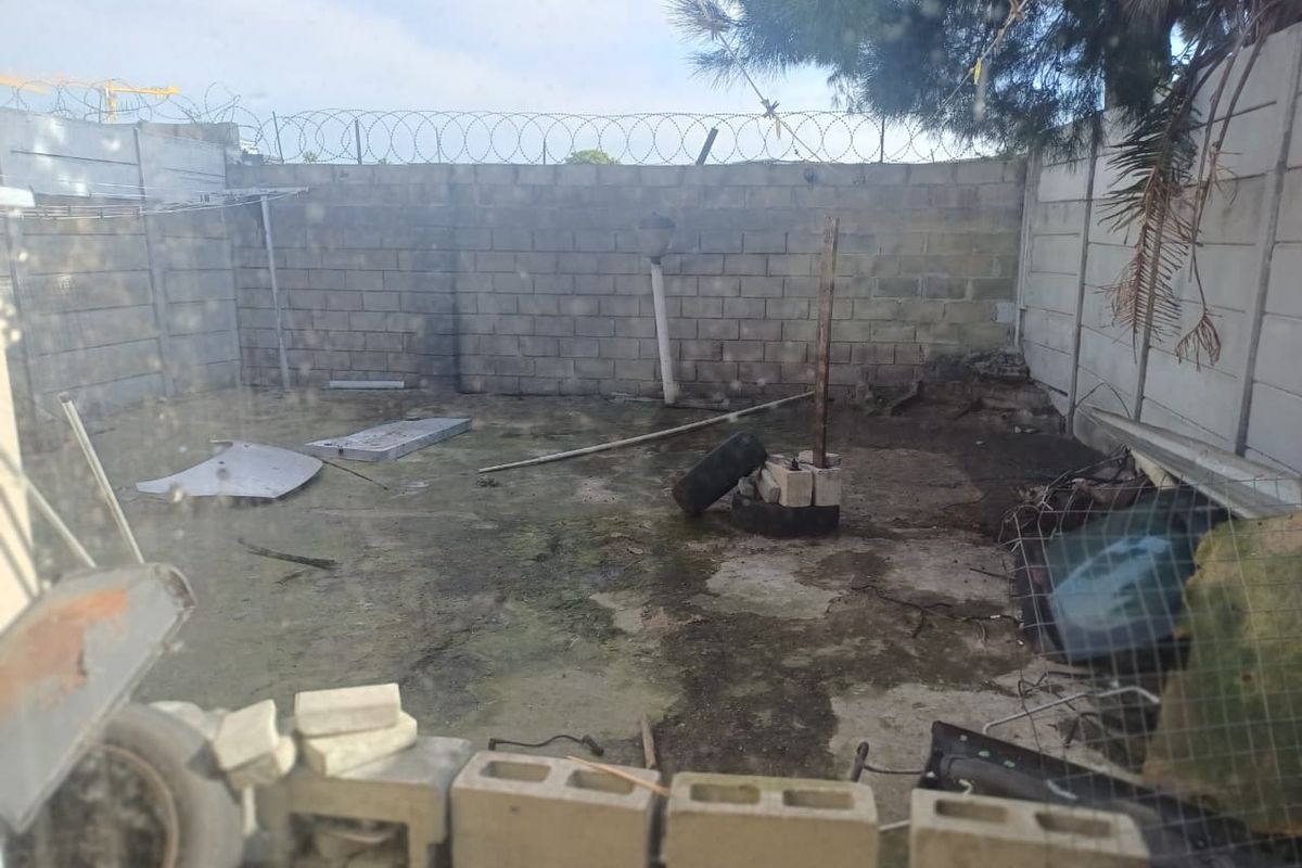 A rear outdoor area featuring concrete block boundary walls with razor wire and paved ground surface in need of maintenance and clearing.