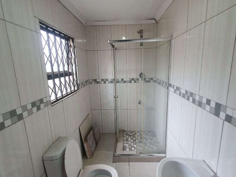 A functional bathroom featuring a shower enclosure, pedestal basin, and toilet, with tiled walls and security bars on the window.