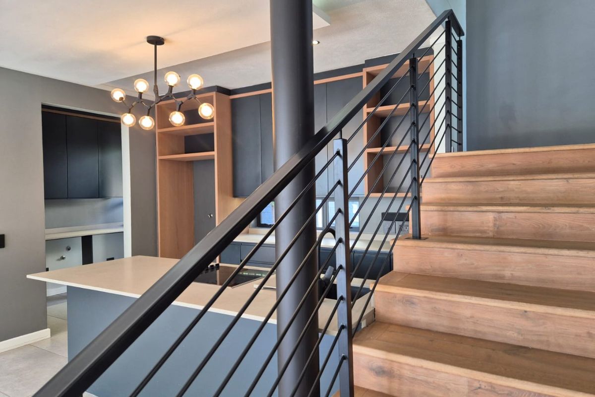 Modern open-plan kitchen featuring dark cabinetry, wood-toned shelving, and a central island, adjacent to a contemporary staircase with a sleek metal balustrade.