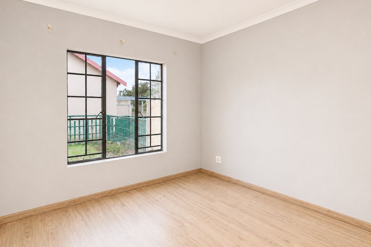 A bright, empty bedroom featuring light-toned laminate flooring, neutral walls, and a black-framed window overlooking the exterior.
