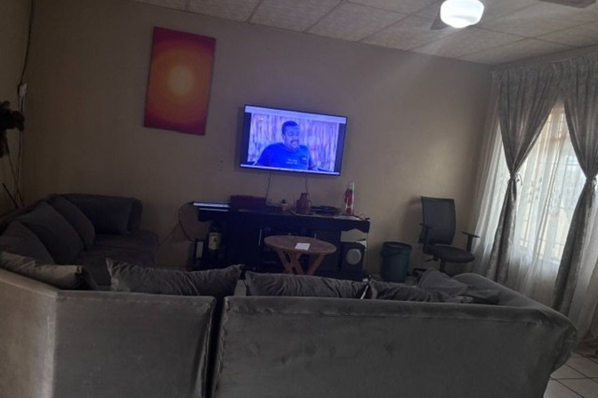 A living room featuring a ceiling fan and tiled flooring, currently showing signs of wear on the drop ceiling tiles.