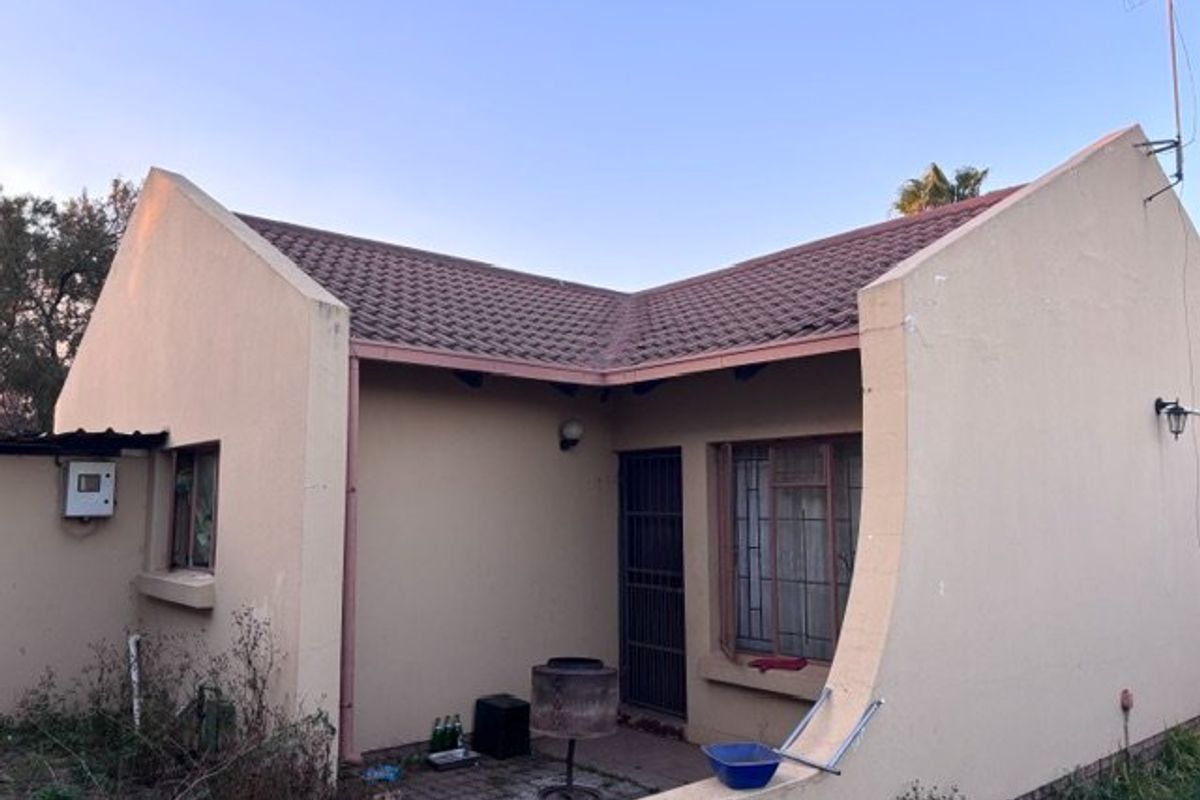 Rear view of the property featuring a tiled roof, security gate, and metal window frames, showing signs of weathered exterior finishes and overgrown garden areas.