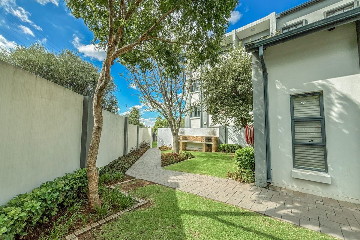 A well-maintained rear garden featuring a paved walkway, manicured lawn, and a built-in brick braai area against the boundary wall.