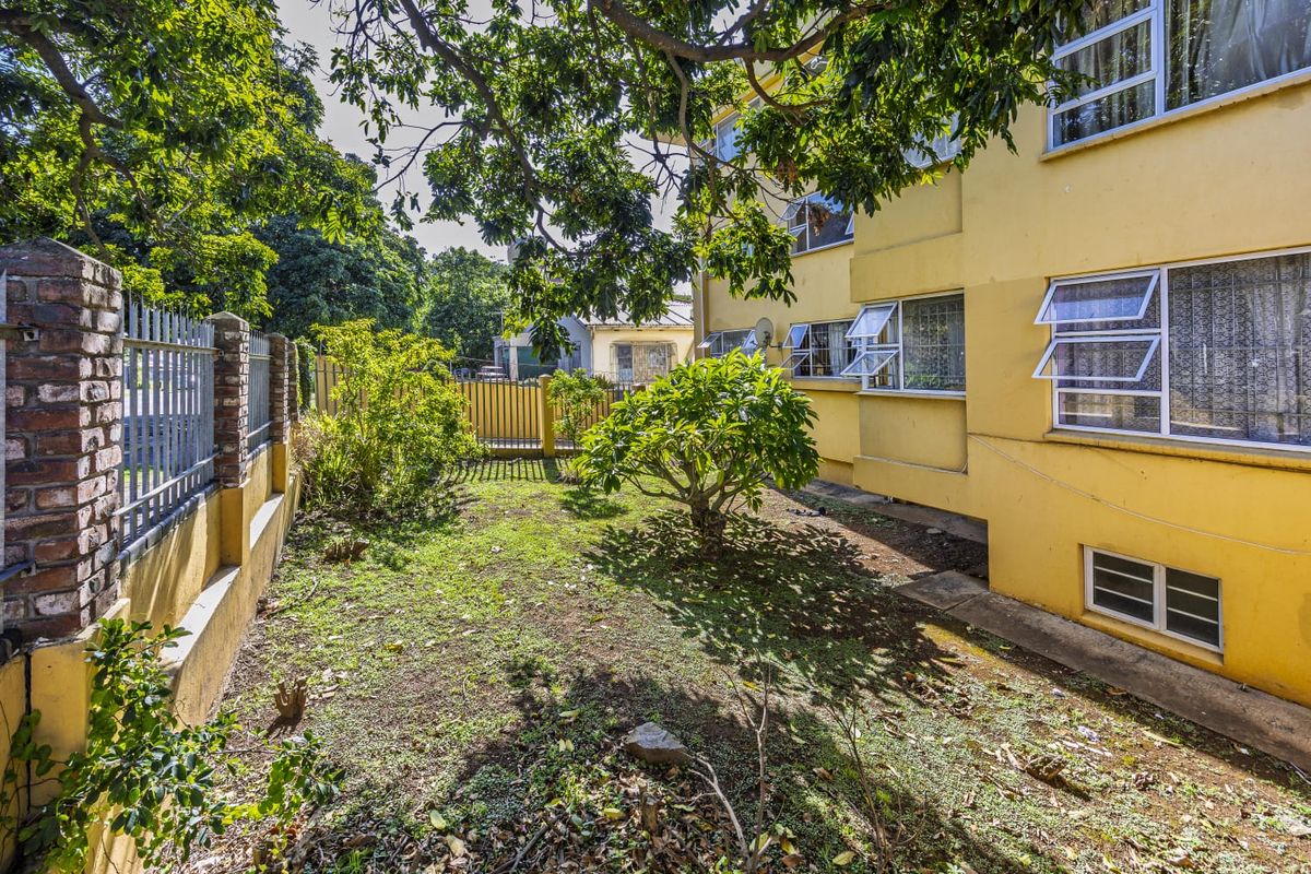 Rear exterior view of the apartment building featuring a small garden area, brick boundary wall, and metal security fencing.