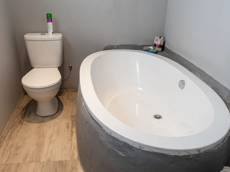A bathroom featuring a built-in oval bathtub with a cement surround, a standard toilet, and wood-look tile flooring.