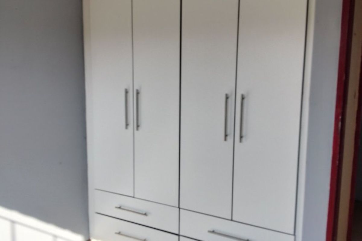 A bedroom featuring white built-in cupboards with modern handles, set against grey walls with visible wear on the flooring and door frame.