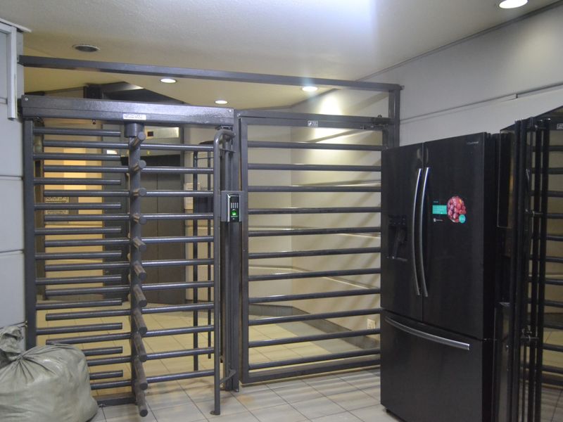 A secure building entrance area featuring a metal turnstile, security gate, and biometric access control system with tiled flooring and recessed lighting.