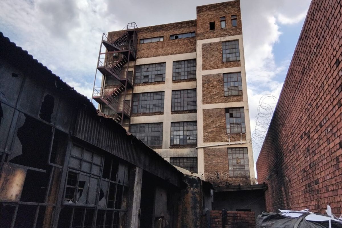 Rear view of a multi-story industrial brick building featuring external fire escapes and significant signs of structural wear and broken glazing.