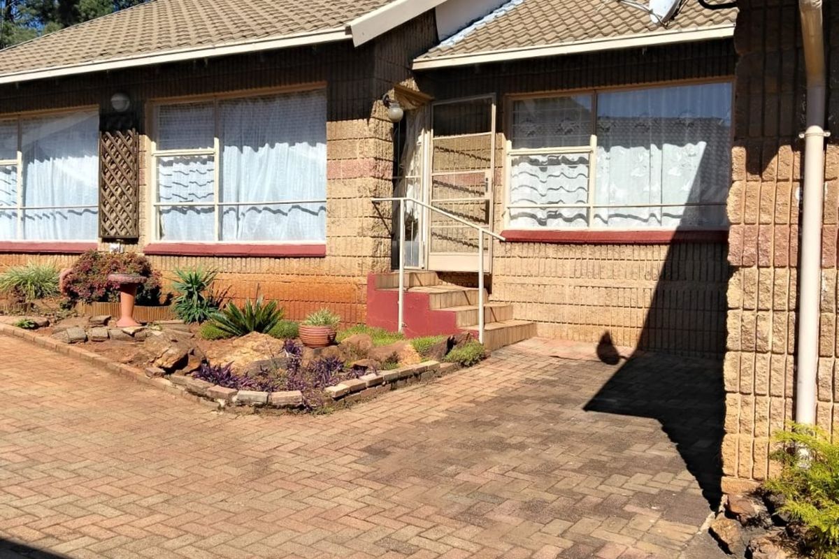 Front elevation of a brick house featuring a paved driveway, tiled roof, and established garden beds with a security gate entrance.