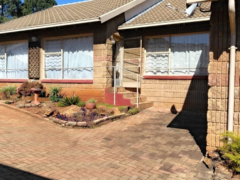 Front elevation of a brick house featuring a paved driveway, tiled roof, and established garden beds with a security gate entrance.