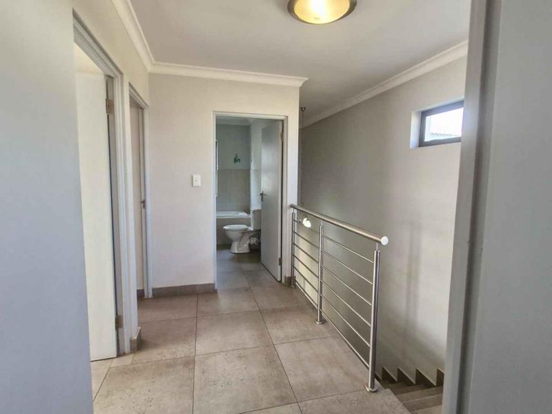 A clean, tiled upper-level hallway featuring a modern stainless steel balustrade overlooking the staircase, with access to an adjacent bathroom.