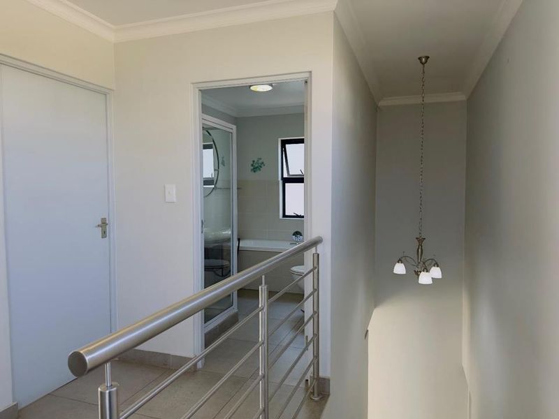 A bright, neutral hallway featuring a modern stainless steel balustrade, tiled flooring, and a classic pendant light fixture, leading into an adjacent bathroom.