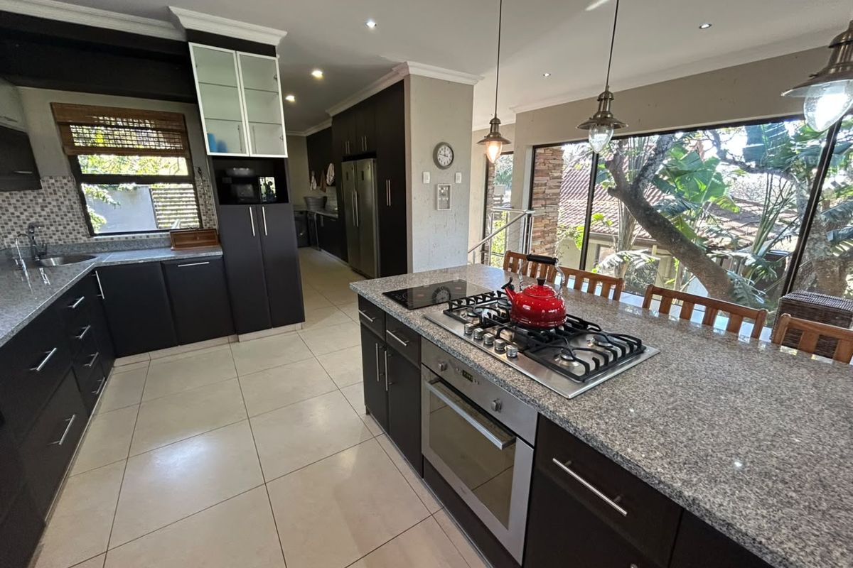 Modern kitchen featuring dark cabinetry, granite countertops, a central island with a gas hob and built-in oven, and large windows overlooking the garden.