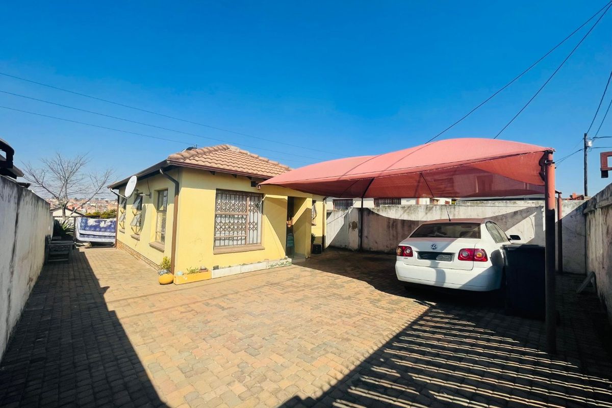 Front exterior view of a single-story house featuring a paved driveway, a shade sail carport, and perimeter boundary walls.