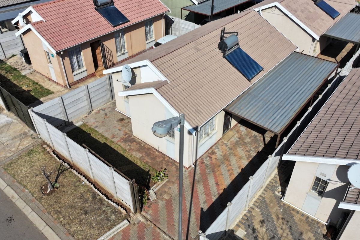 Aerial view of a residential property featuring a tiled roof with a solar water heater, paved driveway, carport, and enclosed boundary walls.