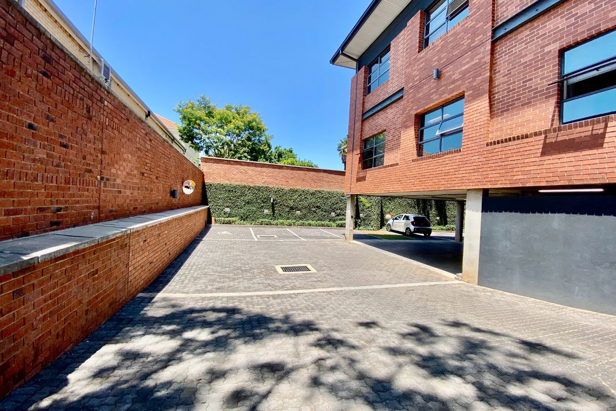 A paved rear exterior area featuring a brick commercial building facade, covered parking space, and secure perimeter walling.