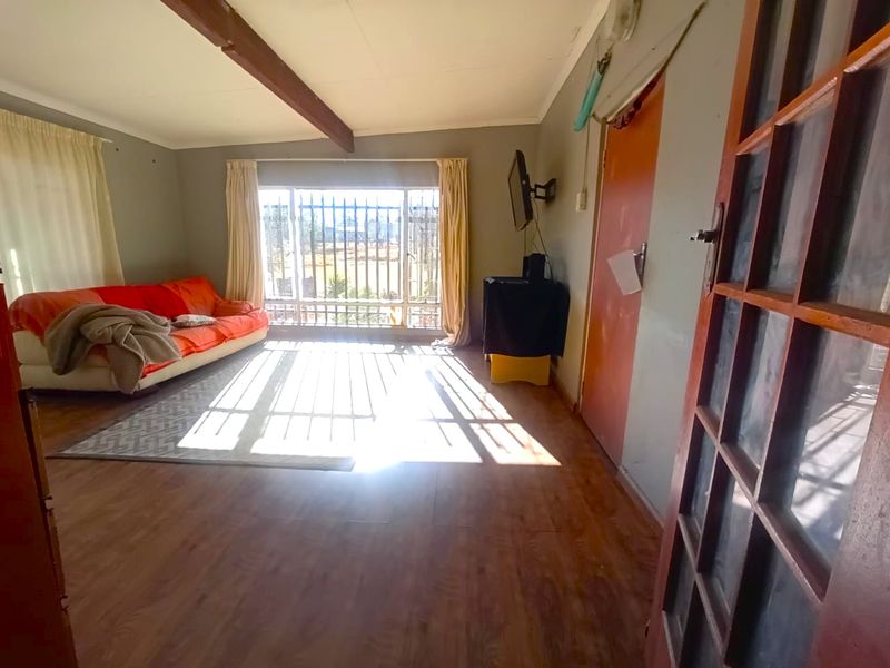 A living room featuring laminate flooring, an exposed ceiling beam, and large windows fitted with security bars, offering natural light.