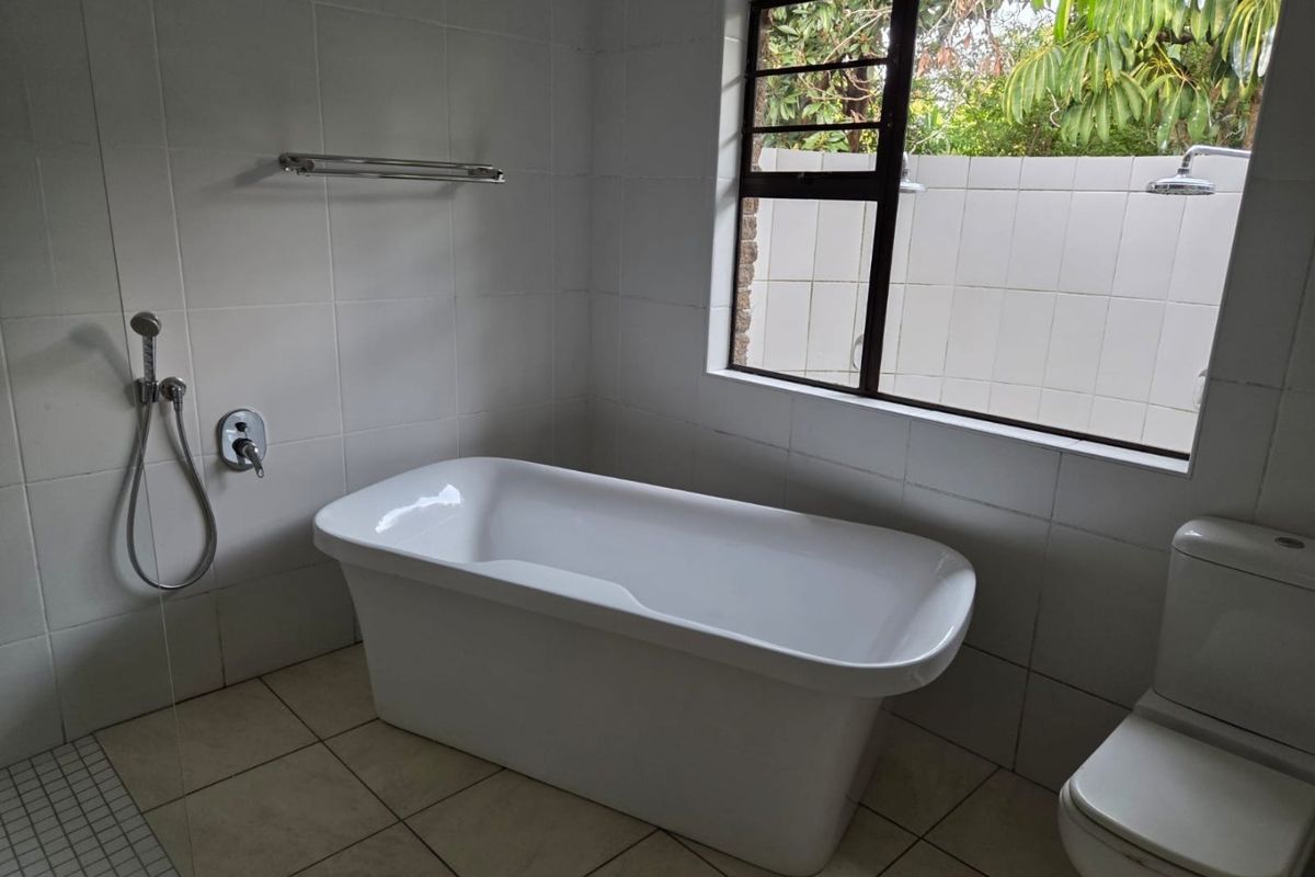 A clean, minimalist bathroom featuring a modern freestanding bathtub, tiled finishes, and a toilet, with natural light provided by a large window.