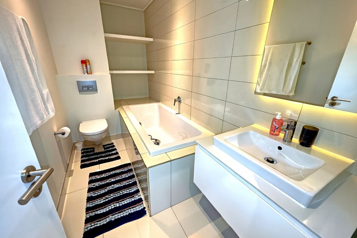 Modern bathroom featuring a floating vanity, built-in bathtub, wall-mounted toilet, and elegant backlit mirror with neutral tiled finishes.