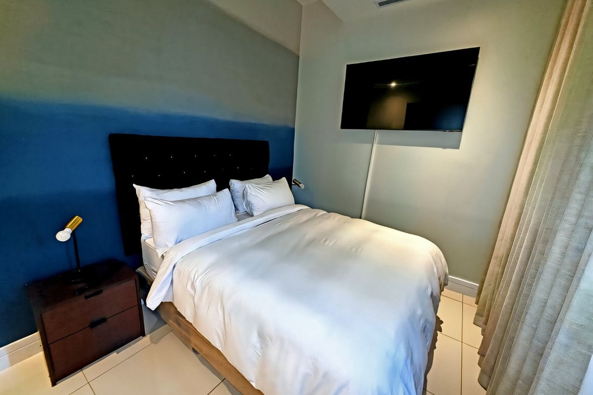 A modern bedroom featuring tiled flooring, recessed ceiling lighting, and a wall-mounted television, finished with a contemporary blue accent wall.