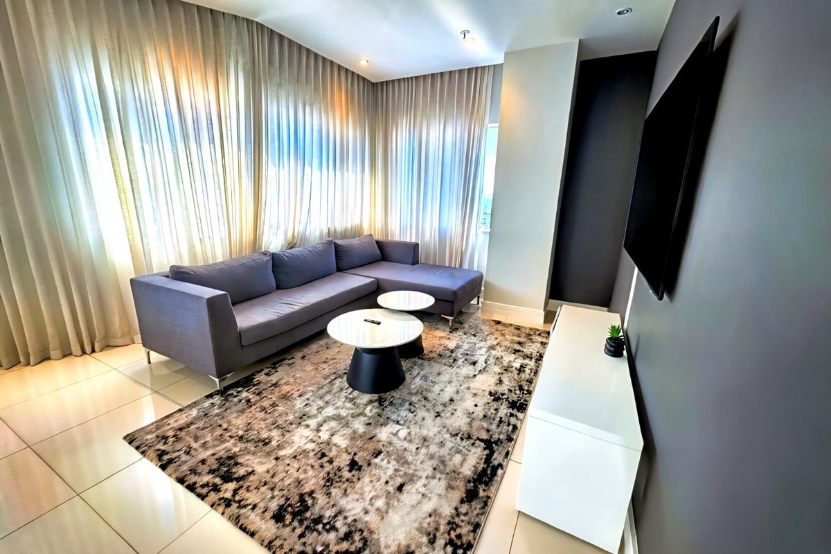 A modern living room featuring tiled flooring, recessed ceiling lighting, and a wall-mounted television, complemented by floor-to-ceiling curtains.