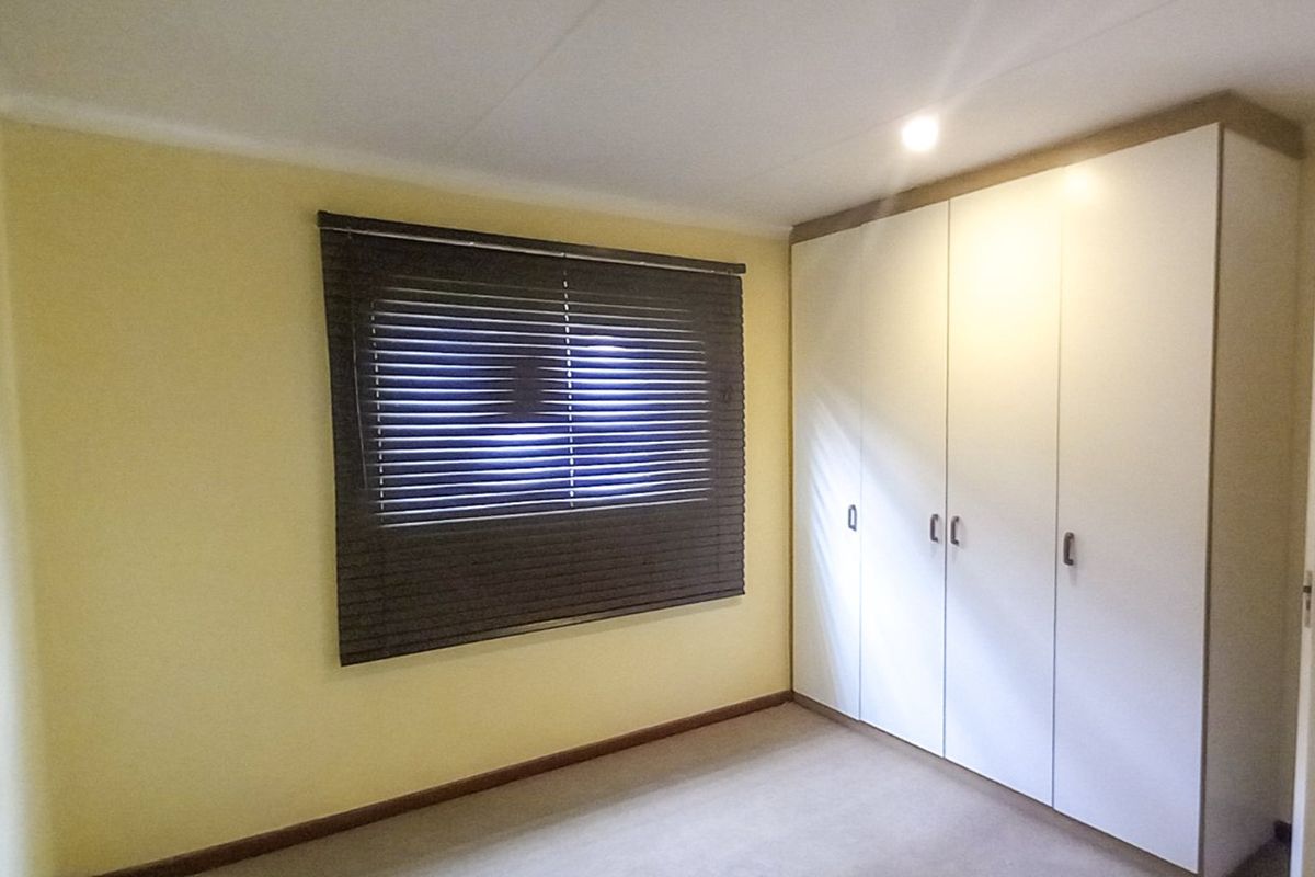 A clean, empty bedroom featuring neutral walls, carpeted flooring, built-in cupboards, and dark venetian blinds.