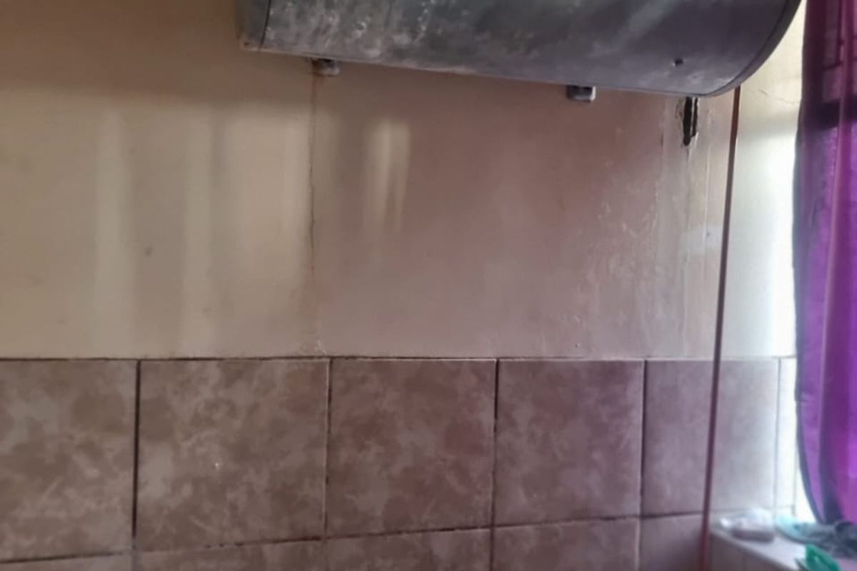 Bathroom featuring a wall-mounted electric water heater, tiled splashback, and bathtub, showing signs of significant wear and maintenance requirements.